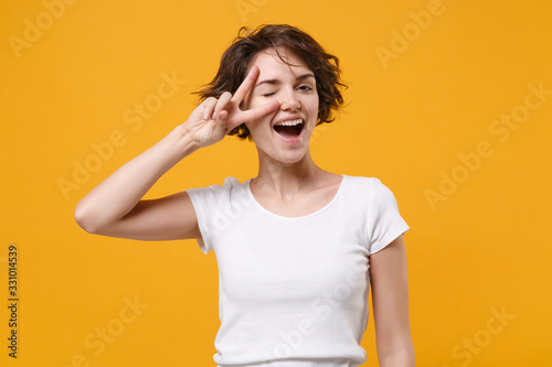 Funny young brunette woman girl in white t-shirt posing isolated on yellow orange background studio portrait. People emotions lifestyle concept. Mock up copy space. Blinking, showing victory sign.