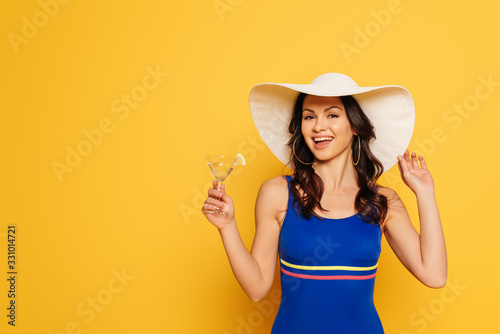 happy woman in swimsuit touching sun hat while holding glass of cocktail isolated on yellow