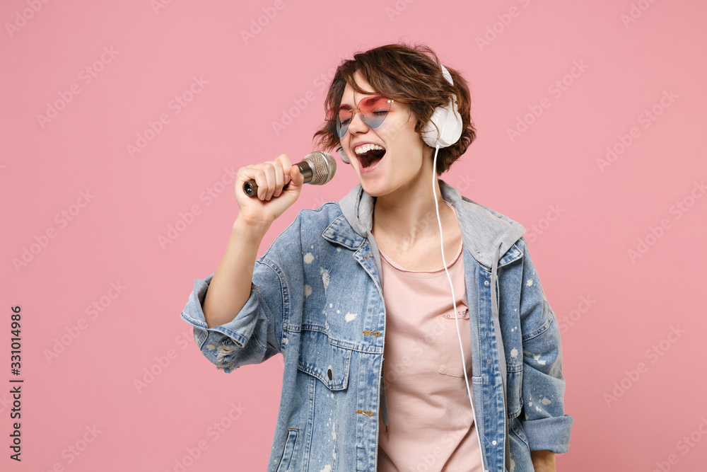 Pretty young brunette woman in casual denim jacket, eyeglasses isolated on pastel pink background. People lifestyle concept. Mock up copy space. Listen music with headphones sing song in microphone. Pretty young brunette woman in casual denim jacket, eyeglasses isolated on pastel pink background. People lifestyle concept. Mock up copy space. Listen music with headphones sing song in microphone.