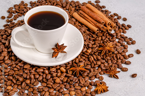 A cup of black aromatic coffee in a white cup with an anise star and cinnamon sticks on a background of roasted arabica coffee beans on a light table
