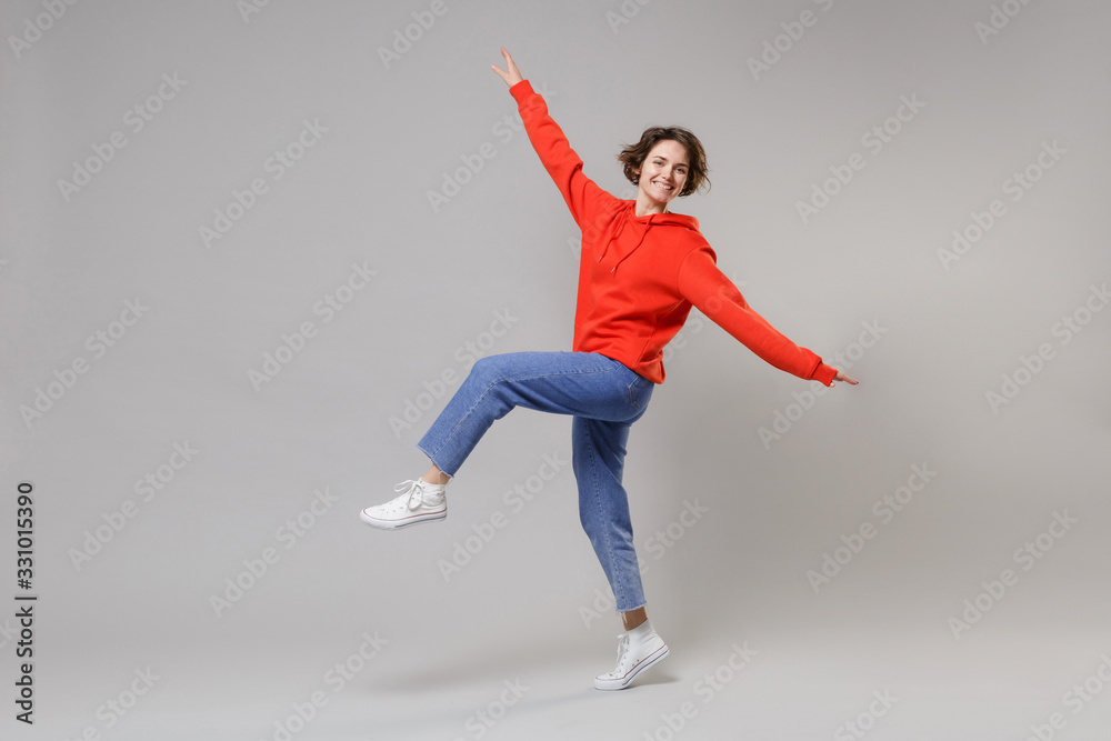 Fototapeta premium Side view of funny young brunette woman girl in casual red hoodie blue jeans posing isolated on grey wall background. People lifestyle concept. Mock up copy space. Dancing, spreading hands and legs.