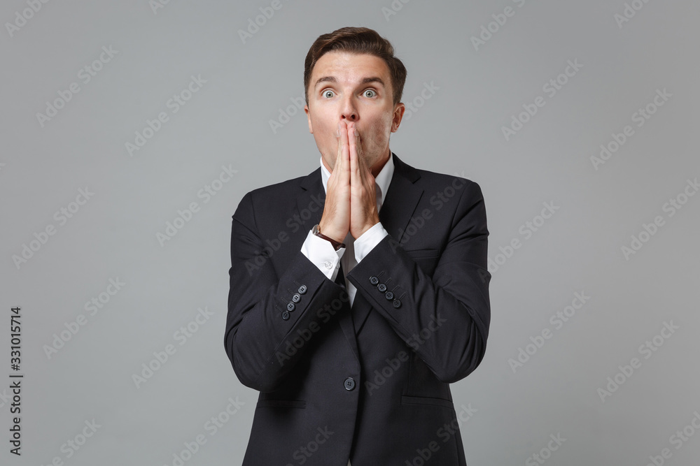 Shocked young business man in classic black suit shirt posing isolated on grey background studio portrait. Achievement career wealth business concept. Mock up copy space. Covering mouth with hands. Shocked young business man in classic black suit shirt posing isolated on grey background studio portrait. Achievement career wealth business concept. Mock up copy space. Covering mouth with hands.