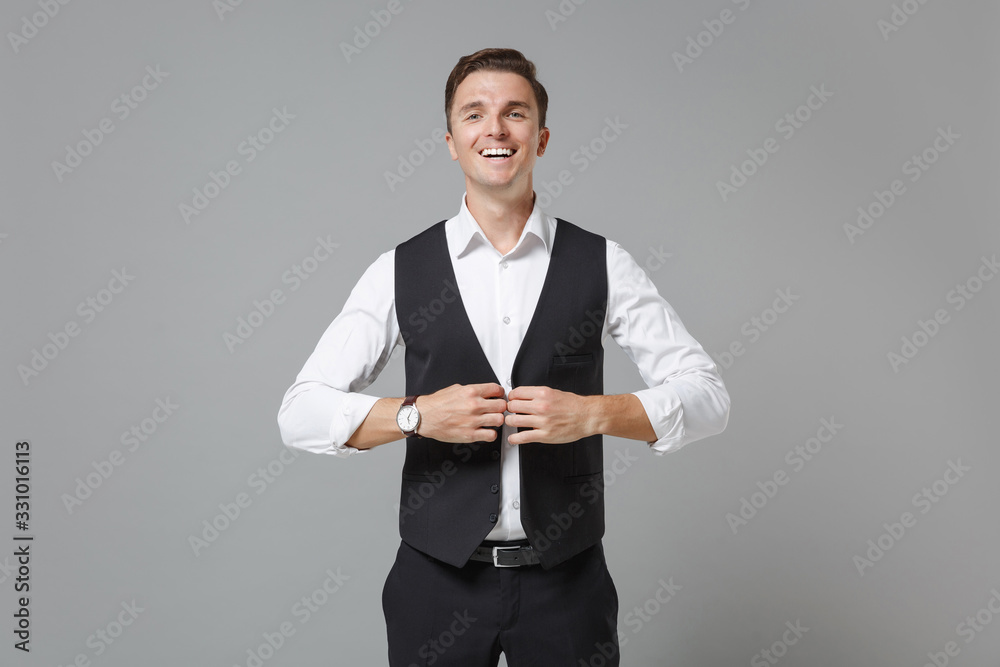 Smiling young business man in classic black waistcoat shirt posing isolated on grey wall background in studio. Achievement career wealth business concept. Mock up copy space. Fastening button on vest. Smiling young business man in classic black waistcoat shirt posing isolated on grey wall background in studio. Achievement career wealth business concept. Mock up copy space. Fastening button on vest.