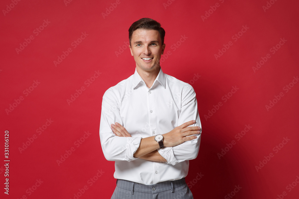 Attractive young business man in white shirt gray pants posing isolated on bright red background studio portrait. Achievement career wealth business concept. Mock up copy space. Holding hands crossed. - obrazy, fototapety, plakaty 