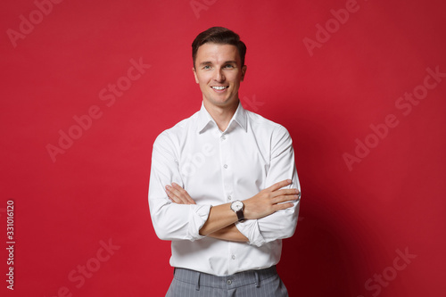 Attractive young business man in white shirt gray pants posing isolated on bright red background studio portrait. Achievement career wealth business concept. Mock up copy space. Holding hands crossed.