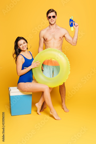 cheerful woman sitting on portable fridge with swim ring near shirtless man holding water gun and on yellow background