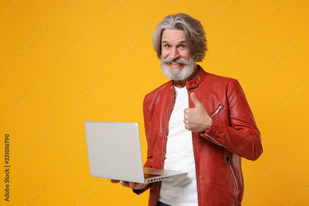Smiling elderly gray-haired mustache bearded man in red leather jacket posing isolated on yellow background. People lifestyle concept. Mock up copy space. Hold laptop pc computer, showing thumb up.