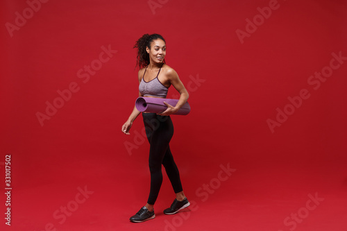 Smiling young african american sports fitness woman in sportswear working out isolated on red background studio portrait. Sport exercises healthy lifestyle concept. Hold fitness mat, looking aside. © ViDi Studio