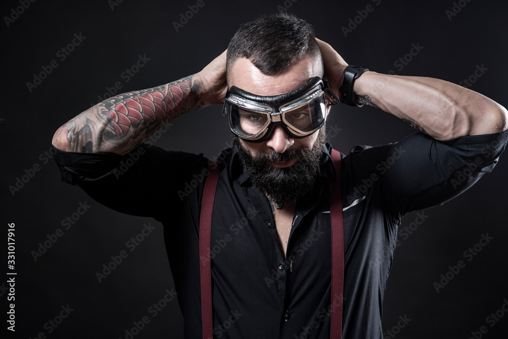 Ragazzo barbuto e Tatuato , vestito con camicia nera e bretelle rosse, si  mette una maschera da motociclista, isolato su sfondo nero Stock Photo |  Adobe Stock