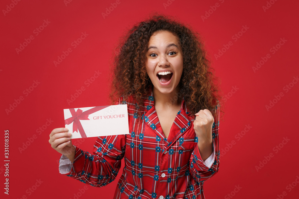 Excited young african american girl in pajamas homewear resting at home isolated on red background. Relax good mood lifestyle concept. Mock up copy space. Hold gift certificate, doing winner gesture. Excited young african american girl in pajamas homewear resting at home isolated on red background. Relax good mood lifestyle concept. Mock up copy space. Hold gift certificate, doing winner gesture.