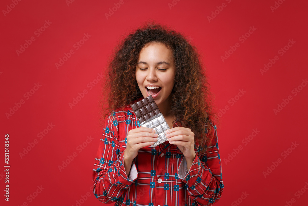 Pretty young african american girl in pajamas homewear rest at home isolated on red background. Relax good mood lifestyle concept. Mock up copy space. Eating biting chocolate bar keeping eyes closed. Pretty young african american girl in pajamas homewear rest at home isolated on red background. Relax good mood lifestyle concept. Mock up copy space. Eating biting chocolate bar keeping eyes closed.