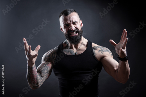 Uomo con barba compeltamente tatuato in canottiera guarda molto irritato con , le braccia e le mani in movimento , isolato su sfondo nero