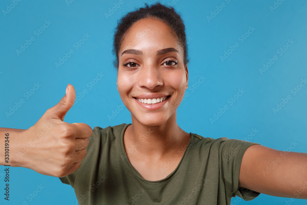 Close up of smiling young african american woman girl in casual t-shirt isolated on blue background. People lifestyle concept. Mock up copy space. Doing selfie shot on mobile phone showing thumb up. Close up of smiling young african american woman girl in casual t-shirt isolated on blue background. People lifestyle concept. Mock up copy space. Doing selfie shot on mobile phone showing thumb up.