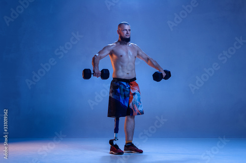Athlete with disabilities or amputee isolated on blue studio background. Professional male sportsman with leg prosthesis training with weights in neon. Disabled sport and overcoming  wellness concept.