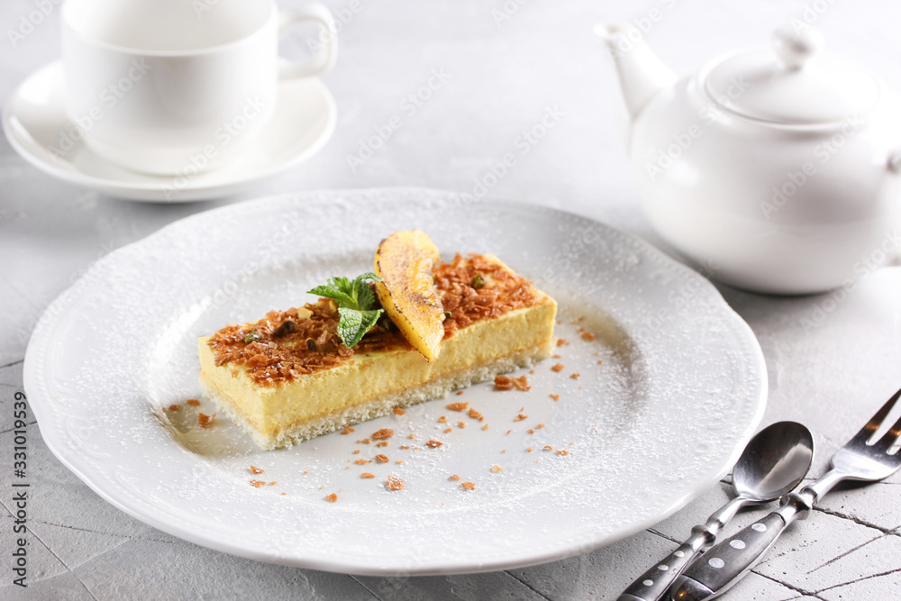 Desserts. Fruit cake with sponge cake and yellow mousse. Mint, yellow slice and powdered sugar on a light plate. Cup6 teapot near plate. Copy space Desserts. Fruit cake with sponge cake and yellow mousse. Mint, yellow slice and powdered sugar on a light plate. Cup6 teapot near plate. Copy space