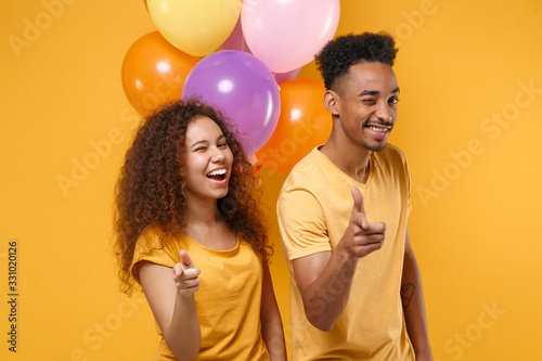 Funny friends couple african american guy girl in casual clothes isolated on yellow background. Birthday holiday party concept. Celebrate hold air balloons pointing index fingers on camera, blinking.
