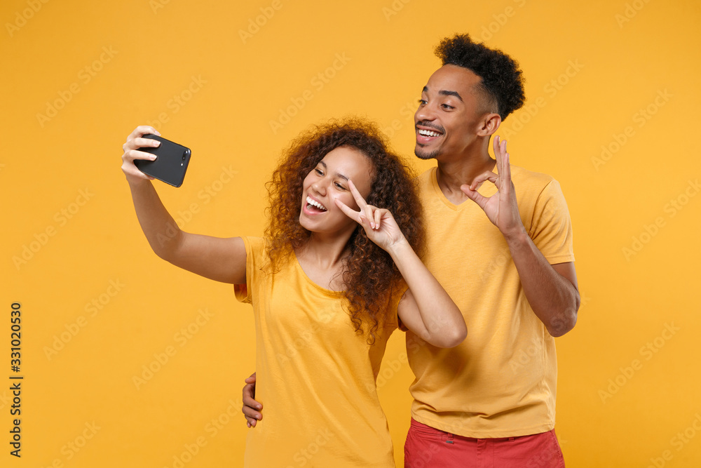 Funny friends couple african american guy girl in casual clothes isolated on yellow background. People emotions lifestyle concept. Doing selfie shot on mobile phone showing OK gesture victory sign.