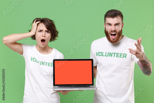 Shocked nervous friends couple in volunteer t-shirt isolated on green background. Voluntary free work assistance help charity grace teamwork. Hold laptop pc computer with blank empty screen screaming.