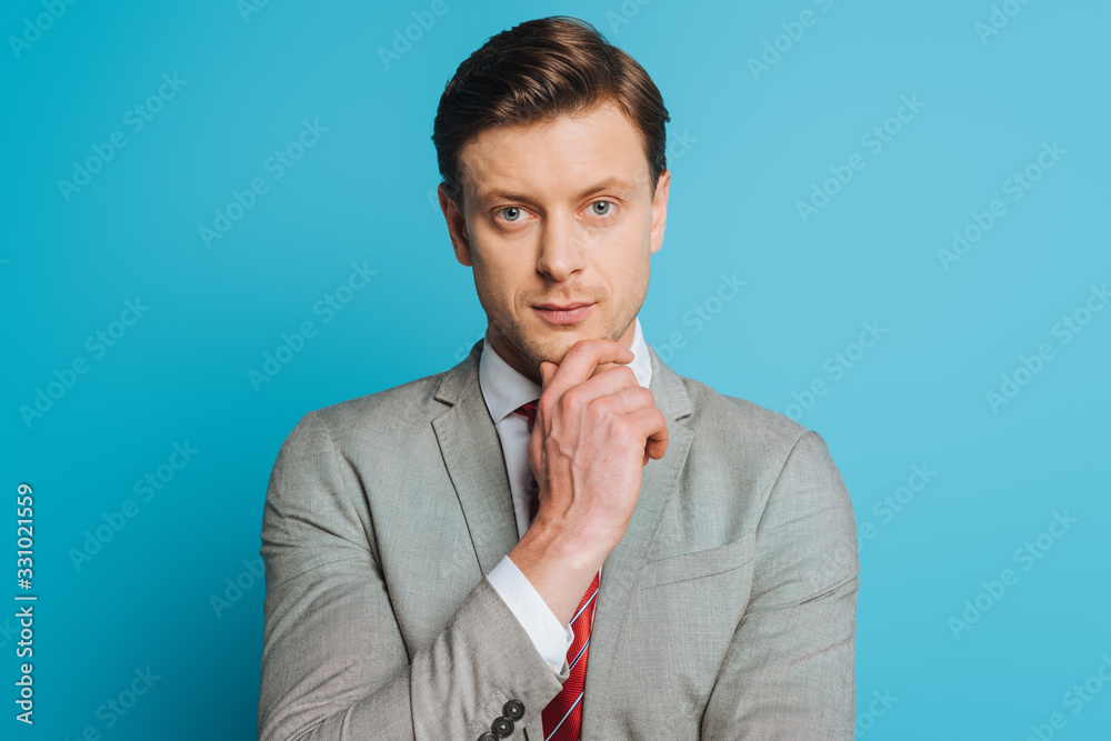serious, thoughtful businessman touching chin while looking at camera on blue background