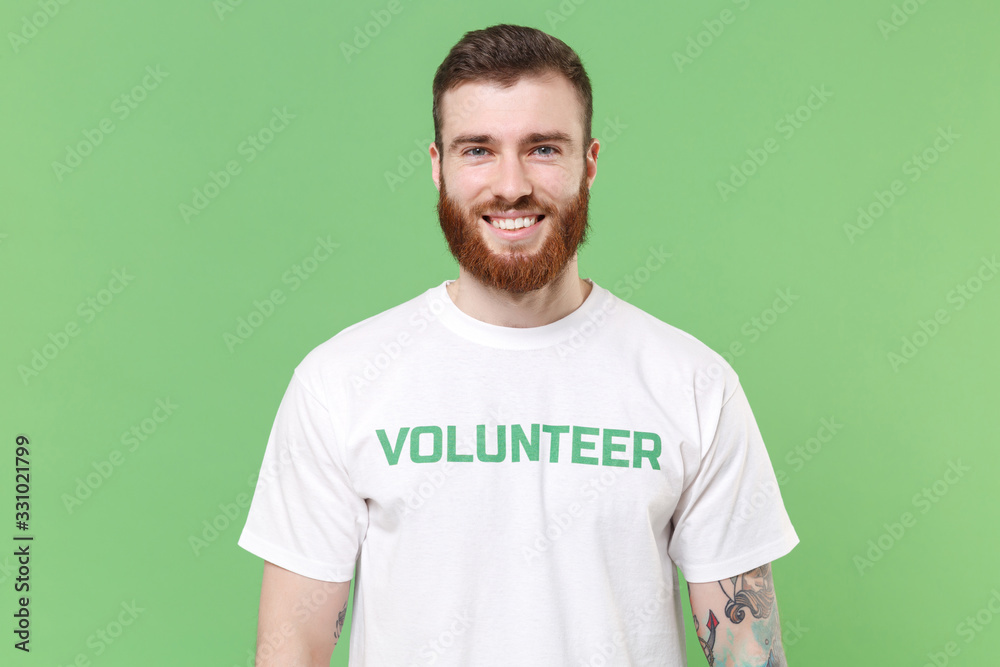 Smiling attractive young bearded man in white volunteer t-shirt isolated on pastel green background studio portrait. Voluntary free work assistance help charity grace teamwork concept. Looking camera. Smiling attractive young bearded man in white volunteer t-shirt isolated on pastel green background studio portrait. Voluntary free work assistance help charity grace teamwork concept. Looking camera.