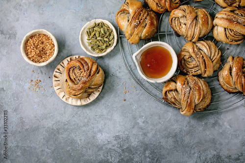 Traditional Swedish cardamom sweet buns Kanelbulle on cooling rack, ingredients in ceramic bowl above over grey concrete texture background. Flat lay, space photo