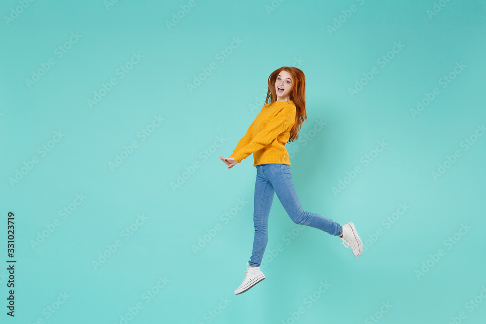 Pretty young redhead woman girl in yellow knitted sweater posing isolated on blue turquoise wall background studio portrait. People emotions lifestyle concept. Mock up copy space. Jumping having fun.