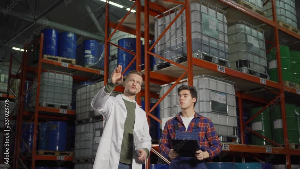 Tracking medium shot of engineer with laptop and young warehouse worker with clipboard standing by steel racks with containers and barrels and discussing work in industrial storage Tracking medium shot of engineer with laptop and young warehouse worker with clipboard standing by steel racks with containers and barrels and discussing work in industrial storage