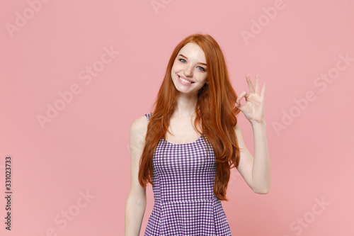 Smiling cute young redhead woman girl in plaid dress posing isolated on pastel pink wall background studio portrait. People sincere emotions lifestyle concept. Mock up copy space. Showing OK gesture.