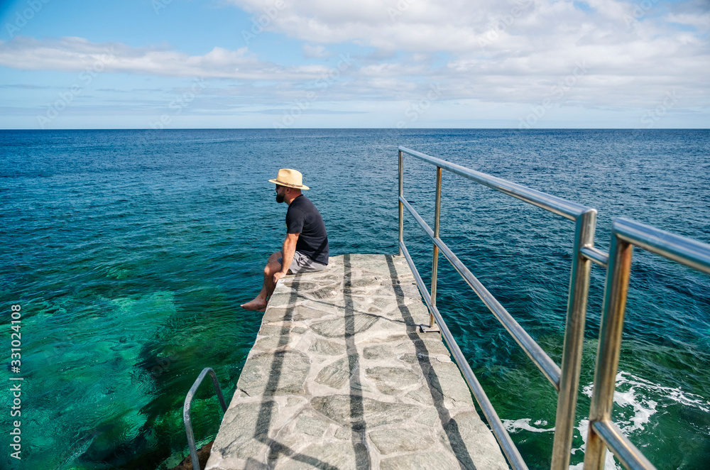 A man with a hat, tourist, in summer, enjoys the view of the sea from a stone walk on the coast
