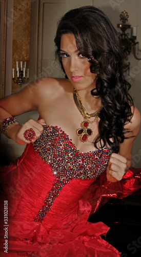 joven quinceañera con maquillaje profesional y cabello arreglado sujetando el corset del traje rojo de pradera con pedrería y pestañas postizas photo