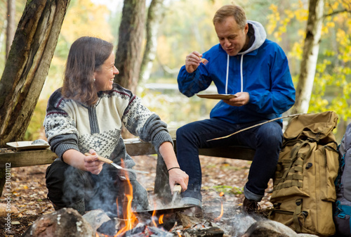 Man and woman making pancakes on campfire in forest on shore of lake, making a fire, grilling. Happy couple exploring Finland. Scandinavian landscape. 