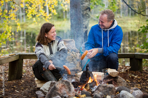 Man and woman roasting on campfire in forest on shore of lake, making a fire, grilling. Happy couple exploring Finland. Scandinavian landscape. 