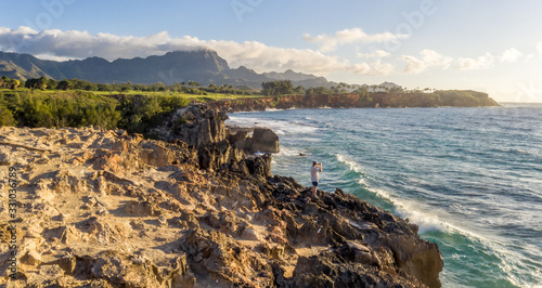 Man standing on a cliff by the ocean watching, looking with binoculars with mountain ridge in the background, Makawehi Lithified Bluff, Kauai photo