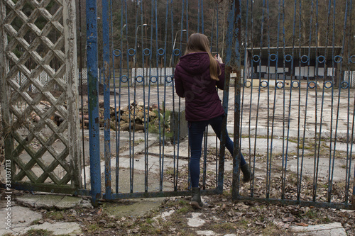 a thin girl with long hair is trying to penetrate through an old blue fence into someone else's private untidy abandoned territory