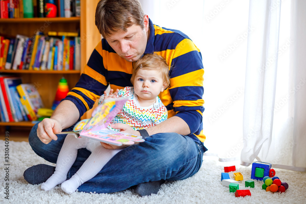 Young father reading book with his cute adorable baby daughter girl. Smiling beautiful child and man sitting together in living room at home. Toddler hearing to dad. Young father reading book with his cute adorable baby daughter girl. Smiling beautiful child and man sitting together in living room at home. Toddler hearing to dad.