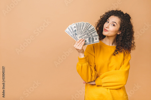 Portrait of a cheerful young woman holding money banknotes and celebrating isolated over beige background.