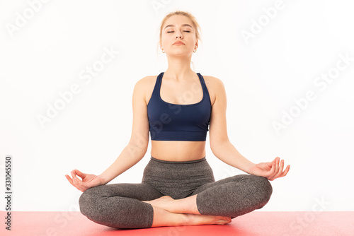 Young attractive sconfident woman practicing yoga, sitting in Half Lotus exercise, Ardha Padmasana pose, working out, wearing sportswear, meditation session, indoor full length