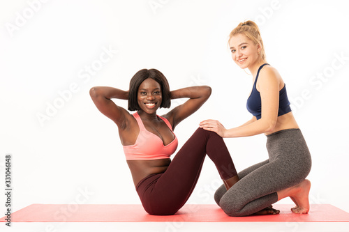 Two young international muscular sportswomen doing pair exercise in sports club. Girlfriends performing sit-ups and push-ups together on mat © Vasya