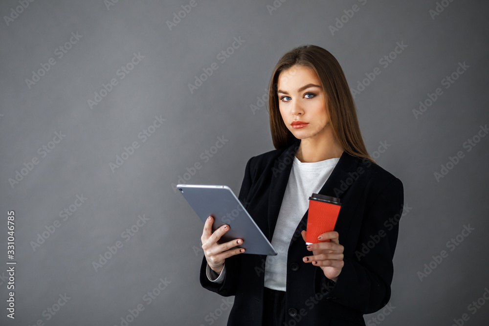 Young pretty girl in business style clothes stands on a gray background with a tablet and a cup of coffee in her hands. Business style Young pretty girl in business style clothes stands on a gray background with a tablet and a cup of coffee in her hands. Business style