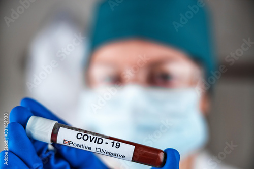 A nurse in a respiratory mask holds a test tube labeled COVID 19, with a positive blood test for a new rapidly spreading coronavirus, close-up, shallow depth of field, selective focus. The concept of