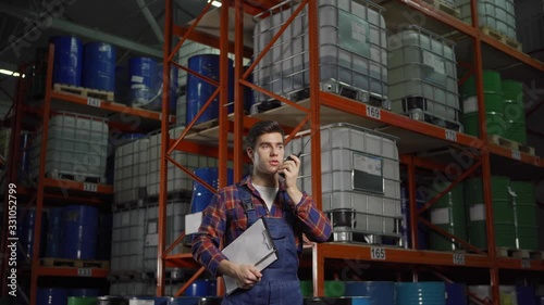 Panning waist up shot of young material handler in uniform talking on walkie-talkie standing by racks with steel barrels and containers in factory storage photo