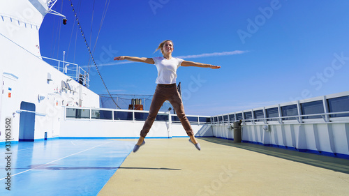 Beautiful sporty girl jumps up on the deck of a cruise ship on a background of blue clear sky. A happy girl smiles on vacation while traveling, jumped high and spread her arms to the sides.