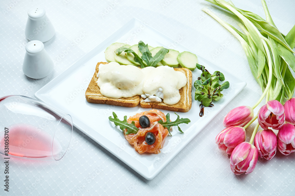 Benedict eggs on toasts with cheese sauce with a side dish of salmon and avocado on a white plate. Restaurant table setting. Close up. Selective focus Benedict eggs on toasts with cheese sauce with a side dish of salmon and avocado on a white plate. Restaurant table setting. Close up. Selective focus