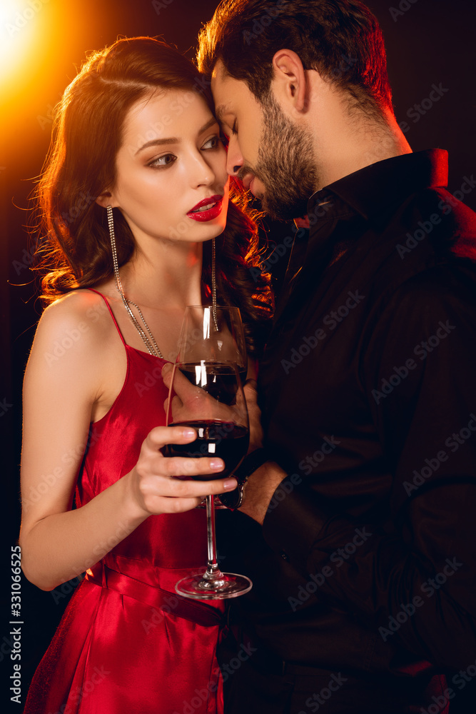 Beautiful woman in dress holding glass of wine near handsome boyfriend on black background with lighting