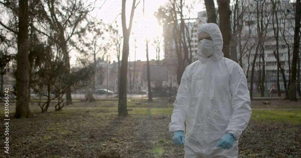 Portrait of male scientist in protective suit wearing a glove and adjusting his mask in daytime park in pandemic coronavirus quarantine time as cars and pedestrians pass by 4K