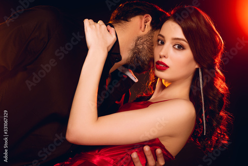 Elegant girl in red dress looking at camera while hugging boyfriend on black background with lighting