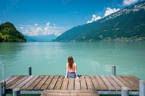 Back view of a young girl sitting on a pier. Beautiful turqouise lake  mountains and sky background. Vacation and traveling concept