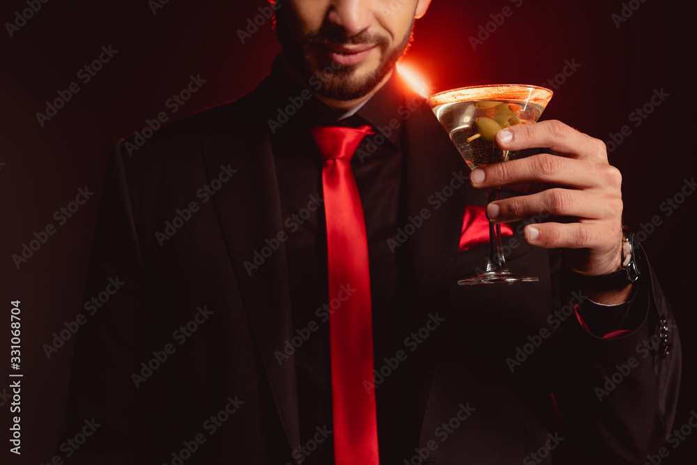 Cropped view of elegant man holding glass of cocktail on black background with lighting