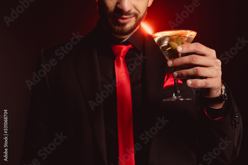 Cropped view of elegant man holding glass of cocktail on black background with lighting