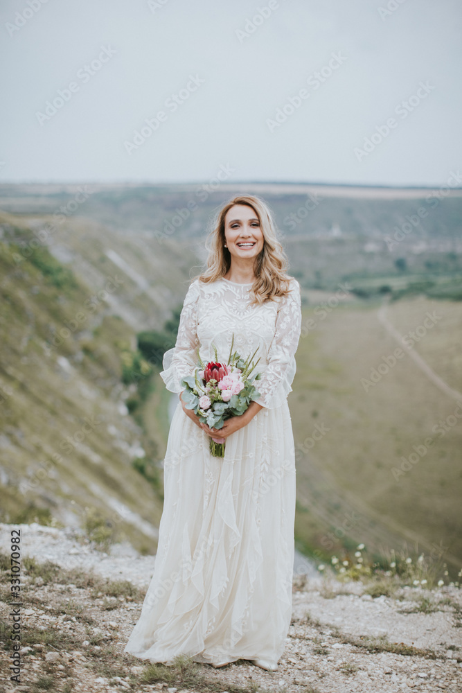 Smiling young blonde caucasian bride in a white, modern lightweight dress posing. Wedding bouquet in her hands. Happy vintage hipster bride, warm colors. Smiling young blonde caucasian bride in a white, modern lightweight dress posing. Wedding bouquet in her hands. Happy vintage hipster bride, warm colors.